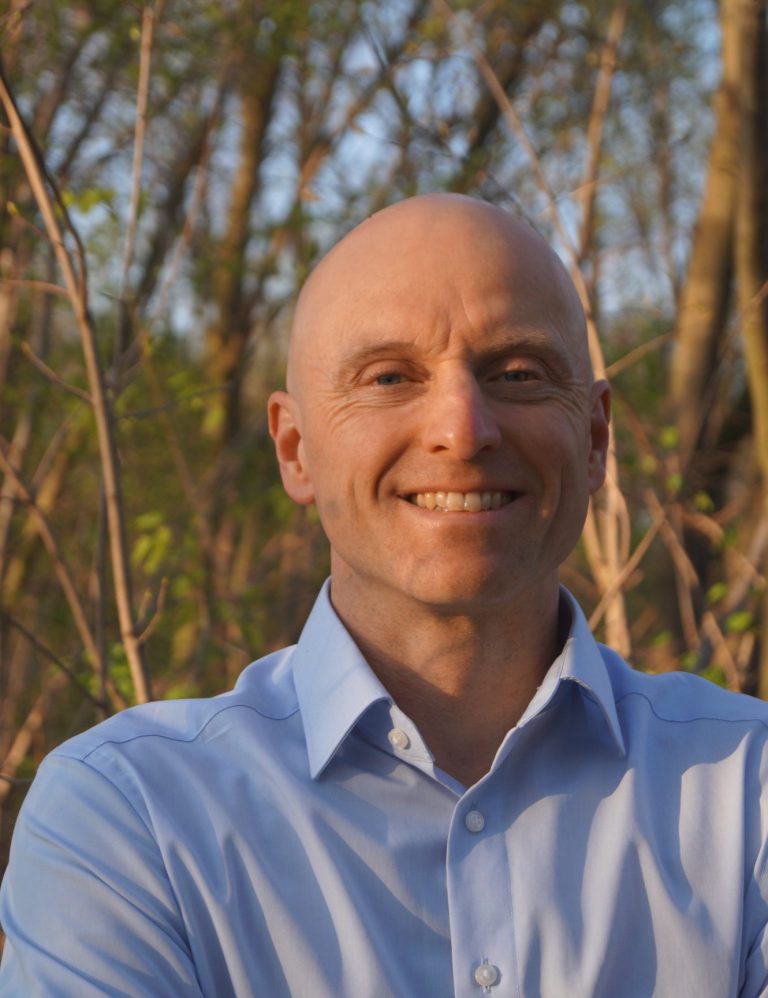Dr. Markus Koschorreck Dr. Markus Koschorreck steht lächelnd vor Bäumen in sonniger Umgebung.